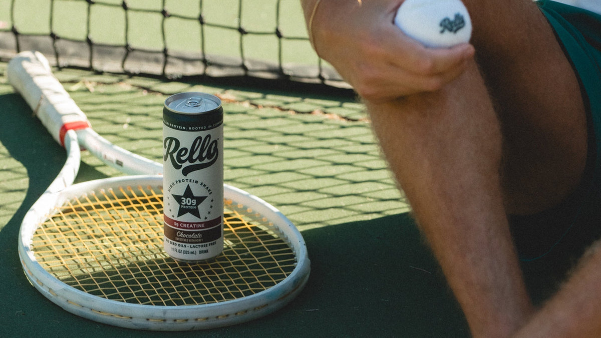 Rello protein drink can sitting on a tennis racket on an outdoor court next to a person holding a tennis ball