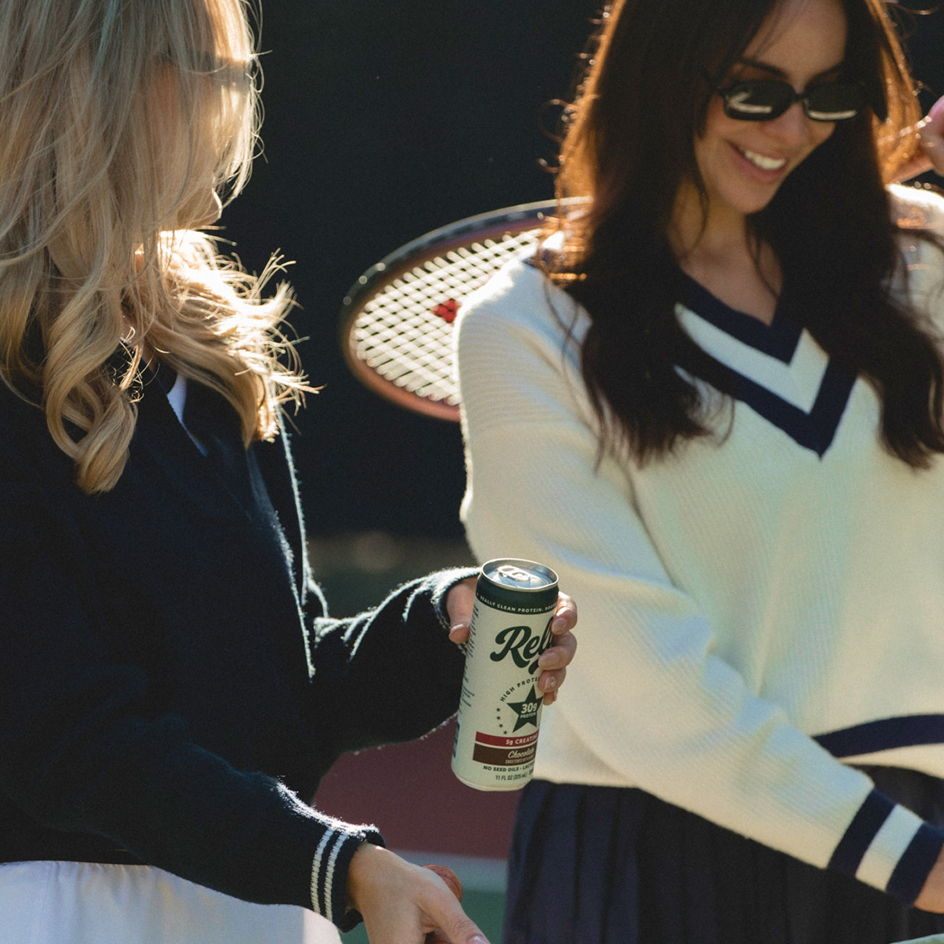 Two women on a tennis court sharing a canned Rello protein drink, one handing the can to the other