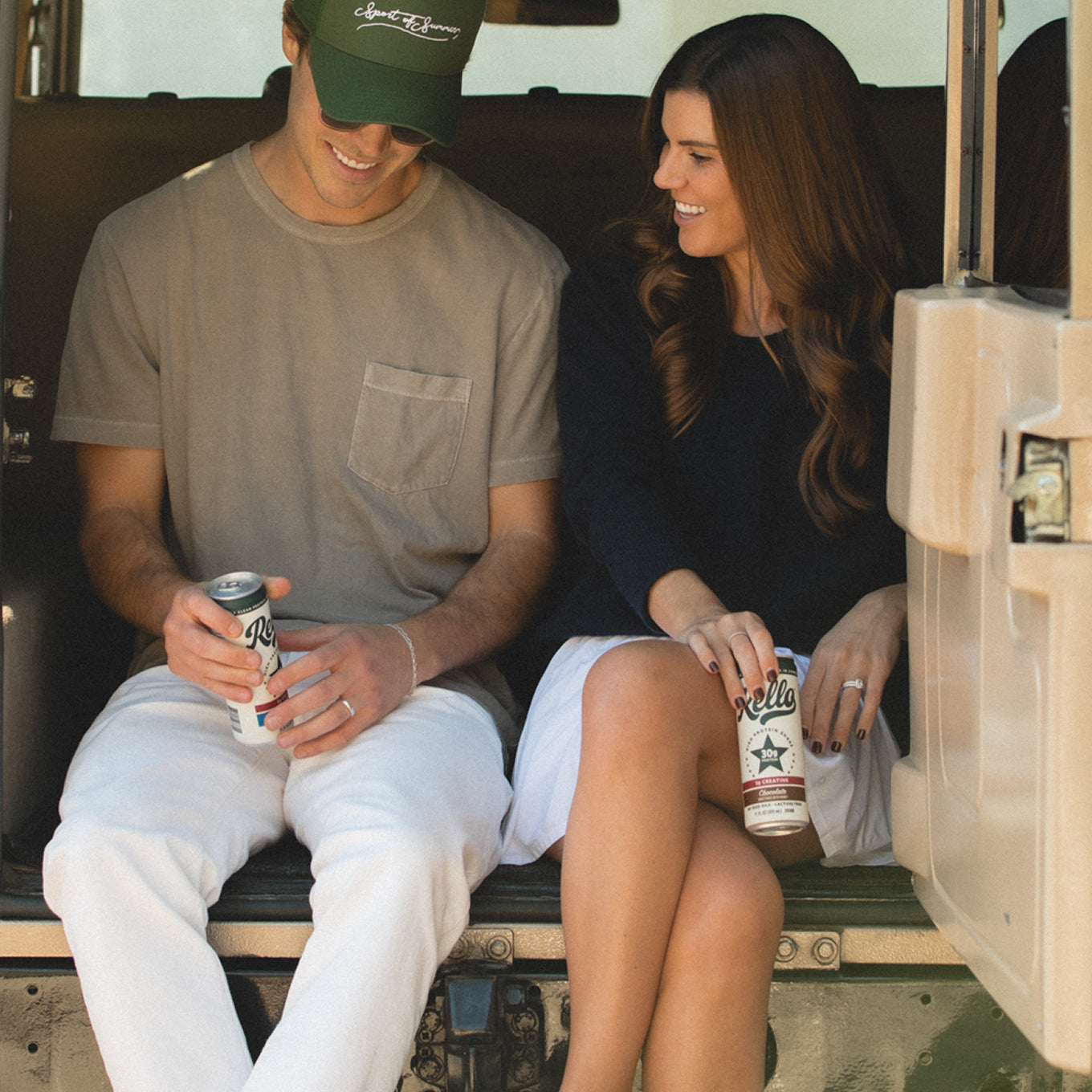 Two people sitting in the back of a vehicle holding Rello protein shake cans and smiling
