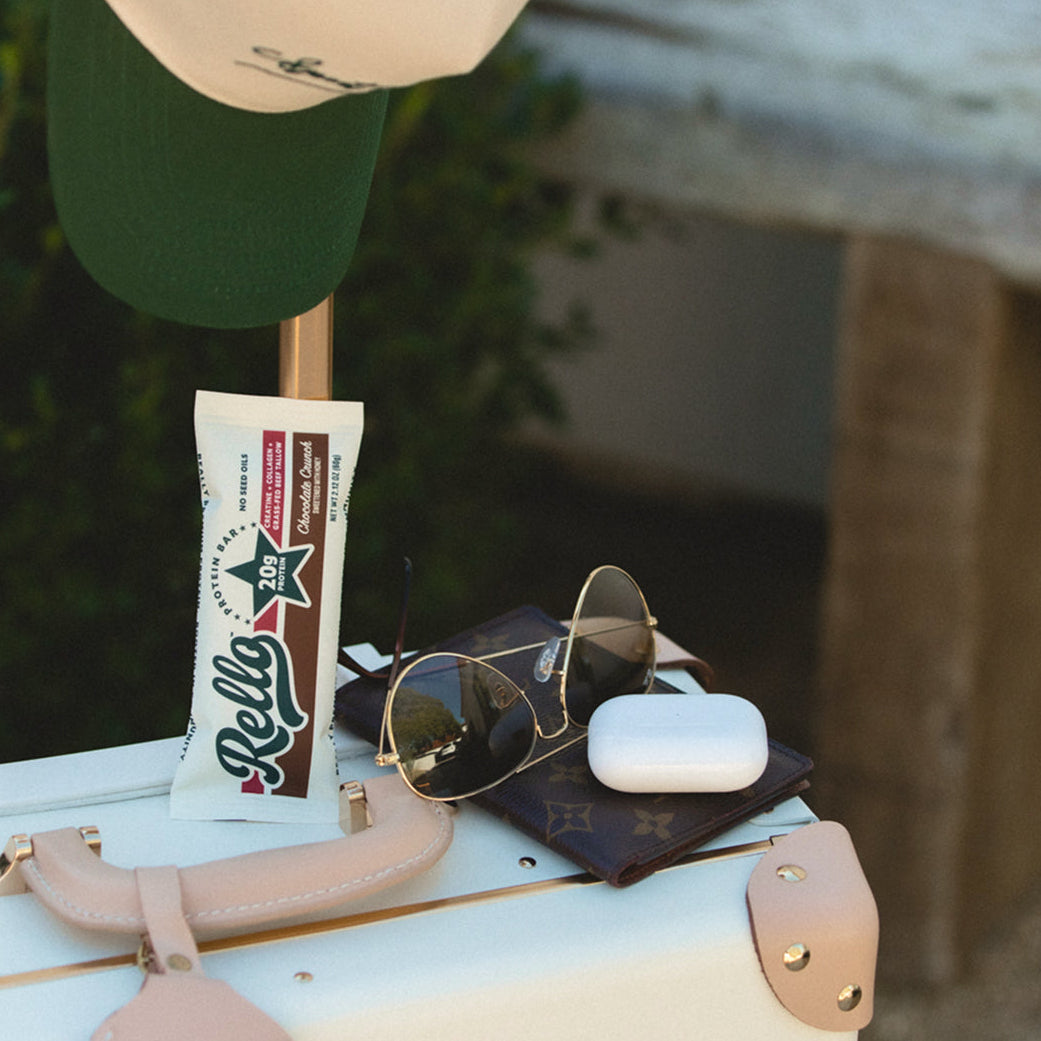 Rello protein bar on a white suitcase with sunglasses, a wallet, AirPods case, and a hat in a travel setting