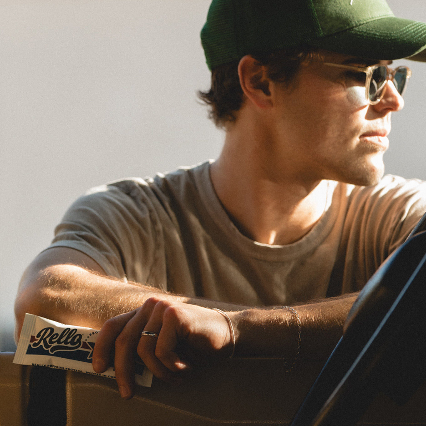 Young man wearing a green cap and sunglasses holds a Rello protein bar while leaning on a car door in warm sunlight
