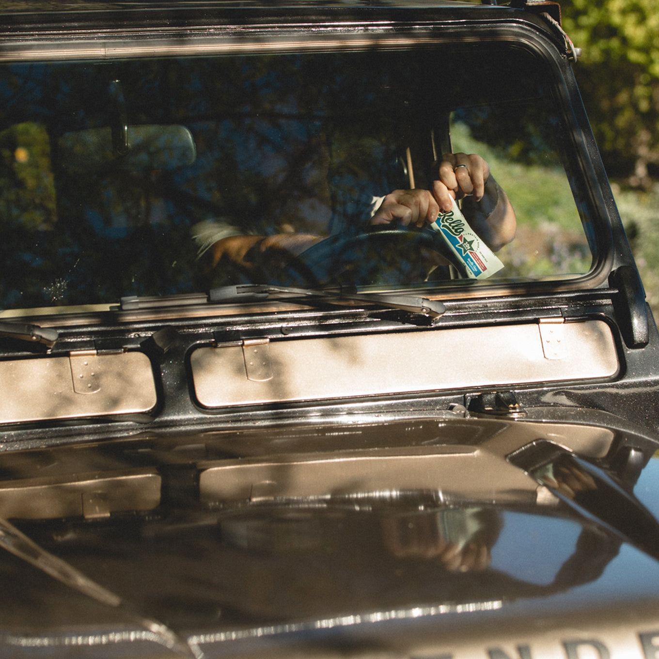 Person in a parked vehicle holding a Rello protein bar up to the windshield in dappled sunlight