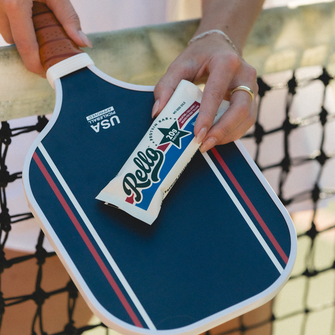 Hands placing a Rello protein bar on a blue pickleball paddle against a court net