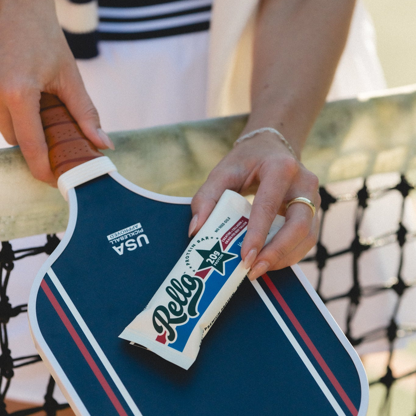 Hands placing a Rello protein bar, wrapper showing Rello logo and 20g protein, on a blue pickleball paddle on a court