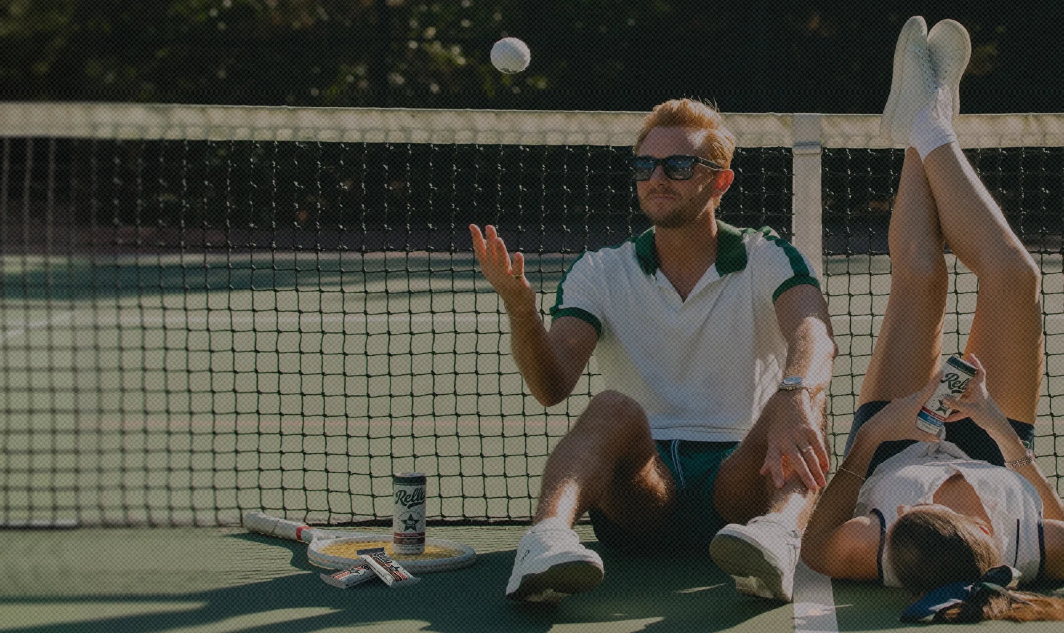 Man tossing a tennis ball and woman lounging on a tennis court, both holding Rello cans with protein bars nearby