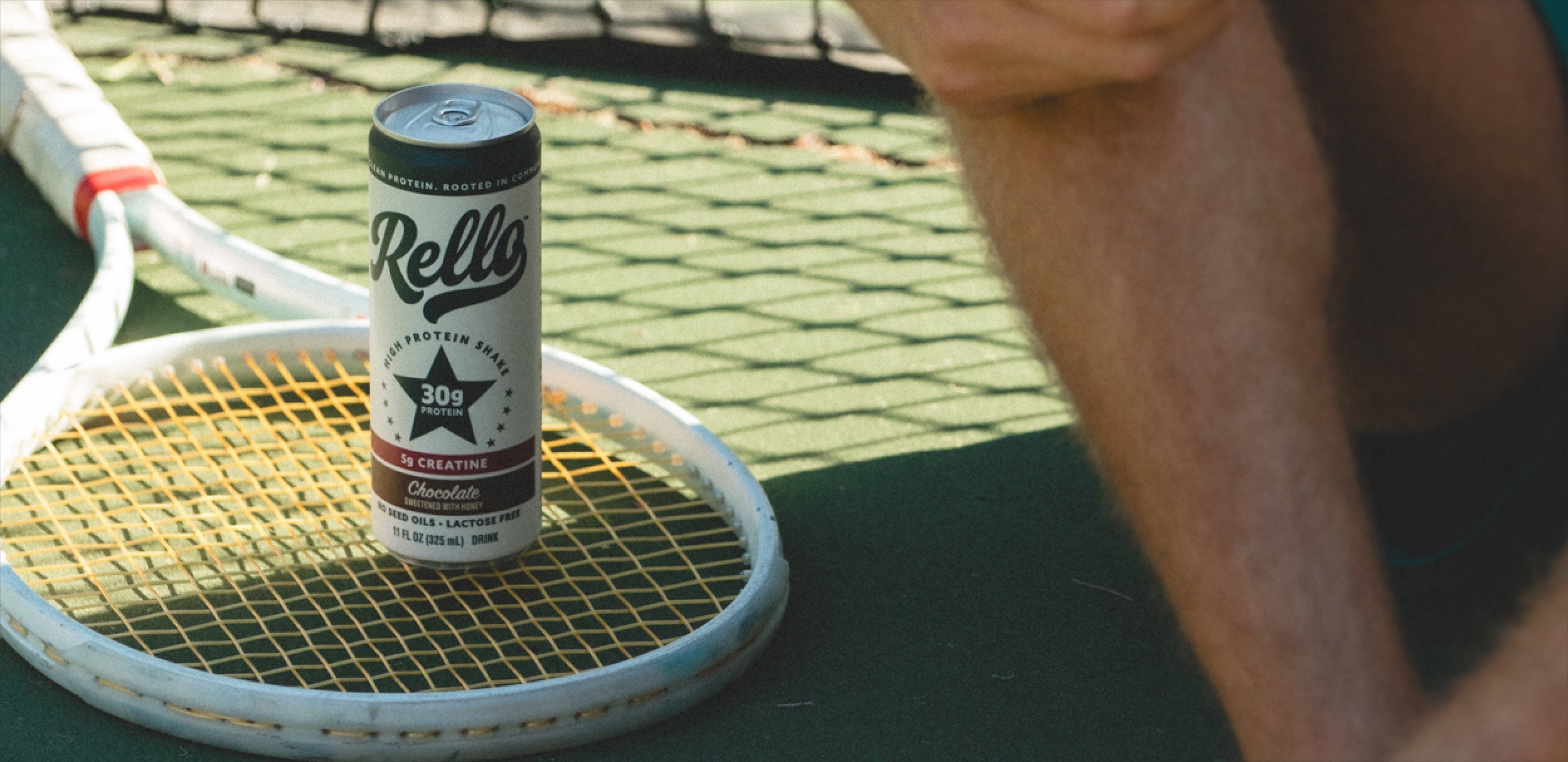 Front view of Rello chocolate protein shake can showing 30g protein and 5g creatine resting on a tennis racket on a court