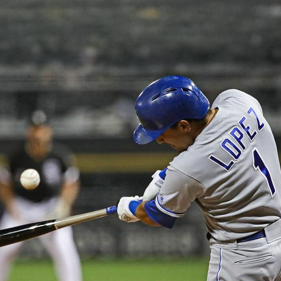 Baseball player swinging a bat during a game on a stadium field, wearing a gray uniform and blue helmet